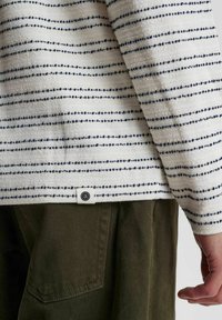 Cămașă din țesătură albă cu dungi orizontale bleumarin și accente punctate; are un mic eticheta cu logo în partea de jos; asociată cu pantaloni de culoare verde măsliniu.