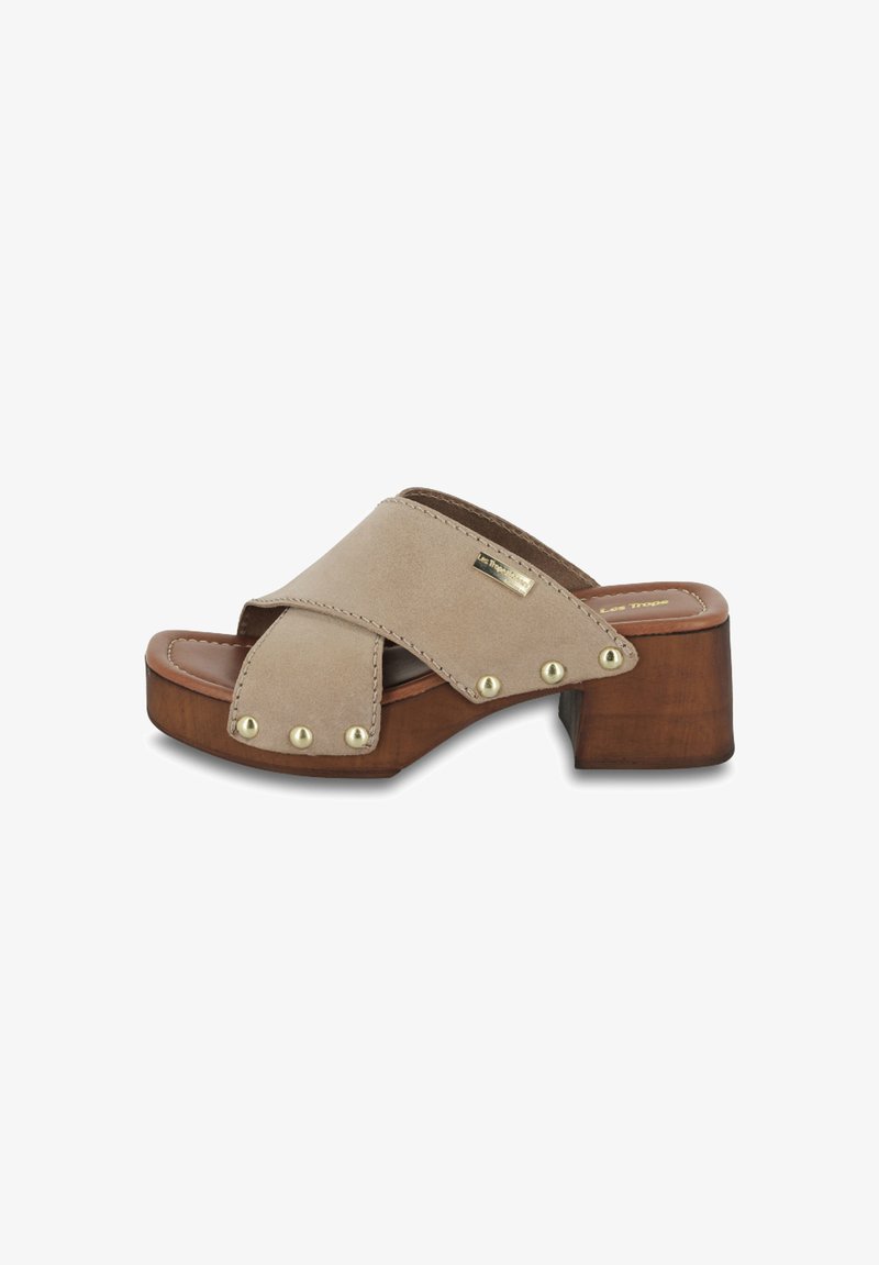 Sandale en suède beige avec un design à lanières croisées, talon en bois et accents en clous métalliques sur les côtés. Texture lisse et forme arrondie.