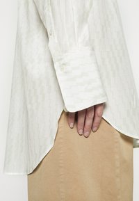 Chemise blanche avec un motif en zigzag vert clair, manches longues, poignet boutonné et ourlet arrondi, associée à une jupe beige. Texture lisse.