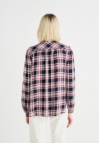 Camicia a maniche lunghe a quadri in blu navy, rosso e bianco. Presenta un colletto, una spalla e un dettaglio di schiena arricciata. Il tessuto è liscio con una leggera lucentezza.