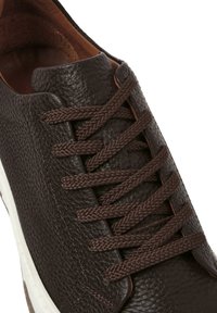 Zapatilla de cuero marrón con superficie texturizada, punta redonda y cordones marrones. Cuenta con una suela de goma blanca y sutiles detalles de costura.