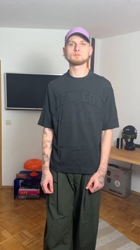 Czarny oversize t-shirt z logo „NEW ERA”, zestawiony z luźnymi oliwkowozielonymi spodniami. Widoczne tatuaże na ramionach.