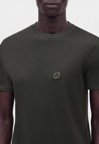 Gros plan d'une personne portant un t-shirt vert foncé à col rond avec un petit logo rond noir et or sur la poitrine gauche.