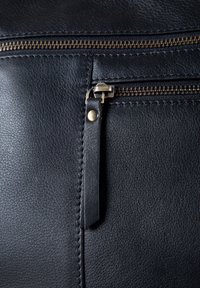 Black leather bag close-up, featuring a visible brass zipper and pull tab, with a textured finish and precise stitching details.