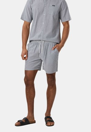 Mann trägt kurzärmeliges, blau-weiß vertikal gestreiftes Hemd mit Knöpfen und passende Shorts mit Kordelzug sowie schwarze offene Sandalen.