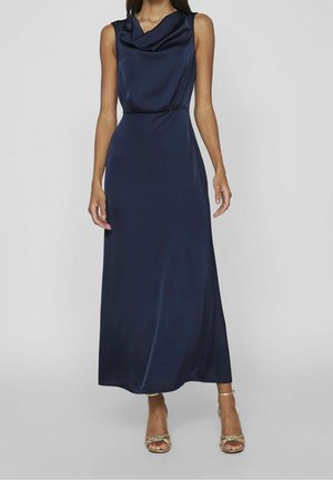 Kobieta ubrana w granatową, bezrękawnikową suknię maxi z jedwabiu o drapowanym dekolcie oraz złote sandały na wysokim obcasie z paseczkami, stojąca na jednolitym tle.