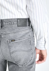 Jeans en denim gris avec un patch en cuir, présentant deux poches arrière et une finition texturée. Portés avec une chemise à rayures bleues.