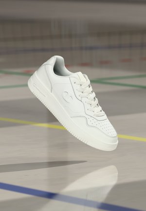 Bílá nízká teniska s hladkým koženým svrškem, perforovanou špičkou a jemnými šedými akcenty. Má ploché tkaničky a strukturovanou podrážku pro lepší přilnavost.