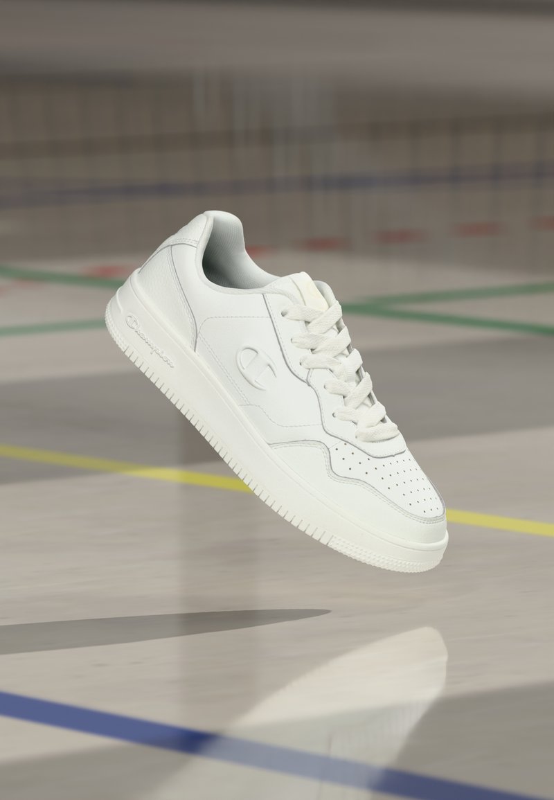 Baskets blanches basses avec dessus en cuir lisse, orteil perforé et accents gris subtils. Dotées de lacets plats et d'une semelle texturée pour une meilleure adhérence.