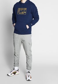 Námořnická mikina s kapucí se žlutým grafickým potiskem, šedé cargo kalhoty s elastickými manžetami a bílé tenisky s černými a červenými prvky.