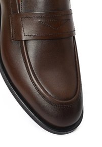 Primer plano de un mocasín de cuero marrón pulido con un detalle de correa cosida y suela negra sobre un fondo blanco.