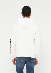 Balts kapučjakas džemperis ar vaļīgu piegriezumu, lielu kapuci un ribotām aprocēm. Piedāvā melnus un sarkānus uzrakstus gar piedurkni, saskaņots ar zilām džinsa biksēm.