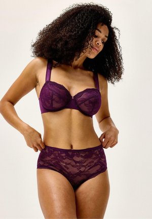 Nainen kiharat hiukset päällä, pukeutunut violetteihin pitsirintaliiveihin ja korkeavyötäröisiin alushousuihin, säätää alushousujen vyötäröä.