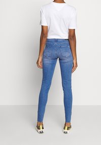 Person som bär en vit kortärmad topp, blå slim-fit jeans med bakfickor och rutiga slip-on sneakers, står med ryggen vänd.