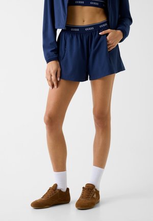 Donna che indossa pantaloncini Guess blu navy e reggiseno sportivo coordinato, calzini bianchi e sneakers marroni, posando con una mano in tasca.