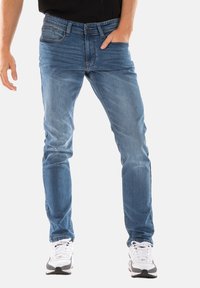 Jean en denim bleu clair coupe slim. Il comporte cinq poches et une fermeture éclair. Porté avec des baskets blanches et noires.