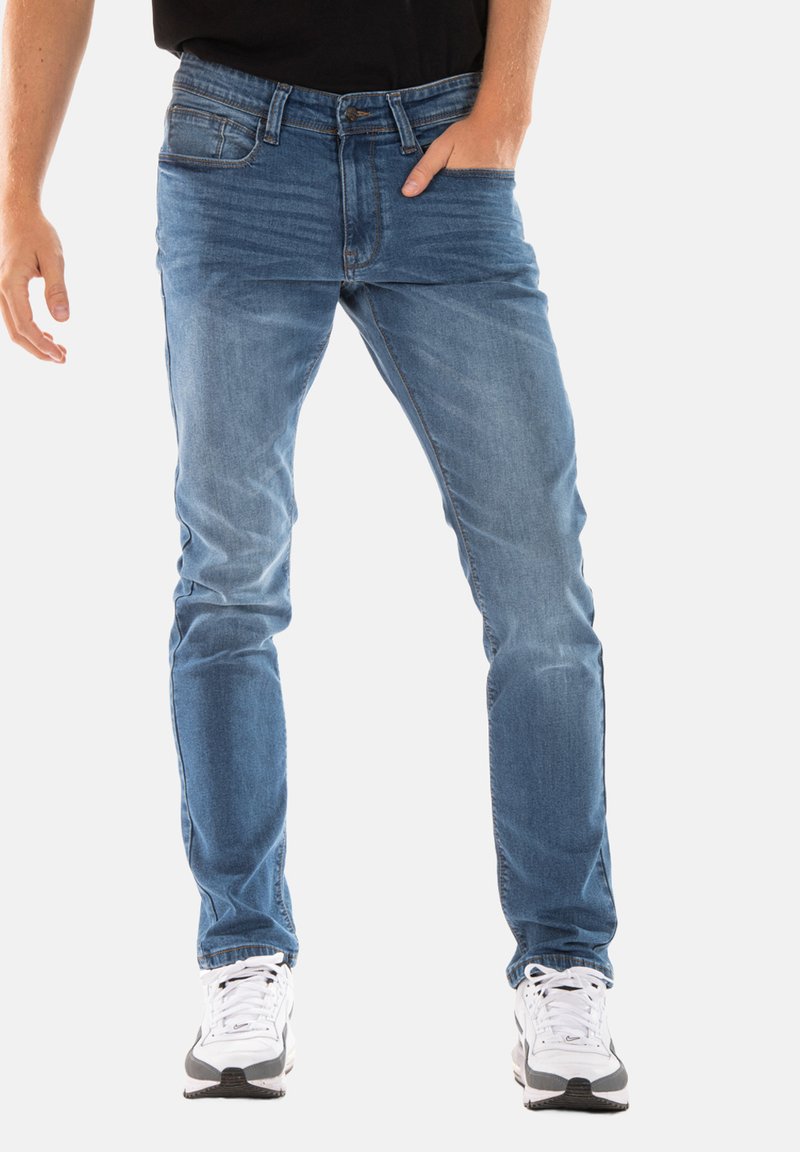 Jean en denim bleu clair coupe slim. Il comporte cinq poches et une fermeture éclair. Porté avec des baskets blanches et noires.