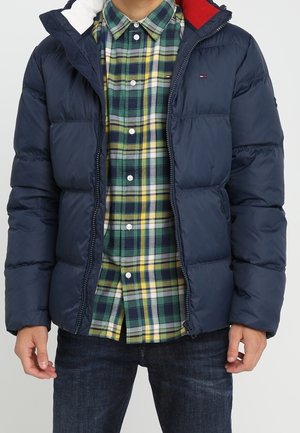 Personne portant une doudoune bleu marine par-dessus une chemise à carreaux verte, jaune et blanche et un jean bleu foncé.