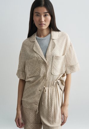 Chemise beige à motif avec des manches courtes, deux poches avant et une taille élastique. Tissu texturé avec un design en grille. Tee-shirt gris visible en dessous.