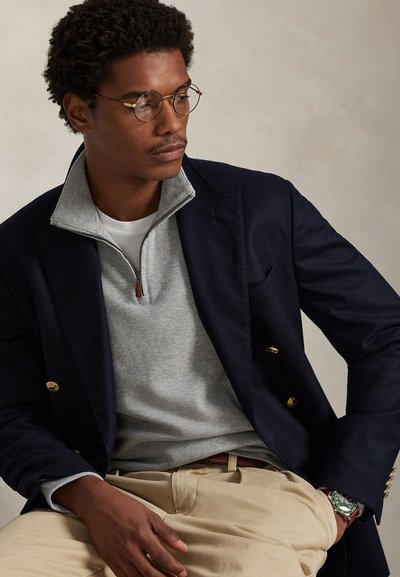 Homme portant des lunettes rondes, un blazer bleu marine avec des boutons dorés, un pull gris à zip court, un pantalon beige et une montre verte au poignet, assis sur un fond neutre.