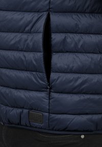 Marineblauwe gewatteerde vest met horizontale quiltpatronen, een ritssluiting aan de voorkant en een gestructureerde stof. Inclusief een stoffen label nabij de zoom.