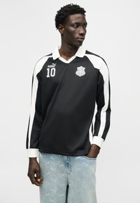 Črno-bela športna majica z dolgimi rokavi z logotipom Puma, številko 10 in grbom, ki jo nosi moški, ki stoji z rokami v žepih.