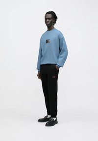 Világoskék pulóver fekete logófolttal, fekete szabadidőnadrággal, melyen egy kis piros részlet található. Az összeállítást fekete cipő egészíti ki.