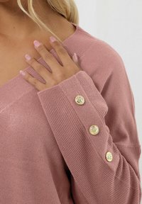 Vaaleanpunainen neulepusero, jossa on syvä V-pääntie, rosoinen pinta ja kultaiset nappikoristeet hihoissa. Pehmeä ja rento malli.