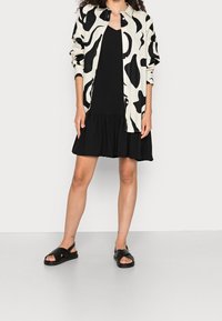 Czarna i biała koszula w wzory, nałożona na czarną sukienkę z rozkloszowanym dołem, w połączeniu z czarnymi sandałami z krzyżującymi się paskami.