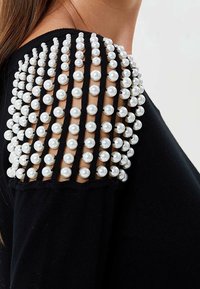 Haut noir avec un design d'épaule composé d'une grille de grandes perles blanches. Tissu doux avec des détails texturés autour de l'encolure.