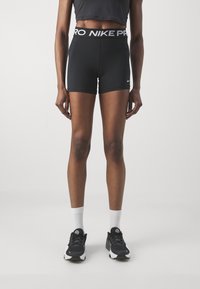 Czarne krótkie spodenki sportowe z szerokim białym paskiem w pasie z napisem "NIKE PRO", dopasowany krój i gładka faktura. Noszone z czarnymi butami i białymi skarpetkami.