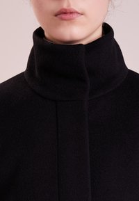 Sort uldfrakke med høj krave og rene linjer. Teksturen fremstår blød og tæt med et minimalistisk design uden synligt metaludstyr.