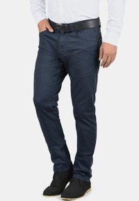 Pantalon slim bleu marine en coton, avec poches avant et ceinture à boucle en cuir noir. Texture lisse, détails minimalistes.