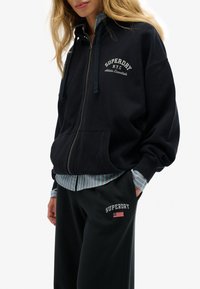 Vrouw in een zwarte Superdry ritssluiting hoodie en bijpassende broek met logo's, gedragen over een gestreept shirt, staande met één hand in haar zak.