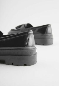 Paire de mocassins en cuir noir avec semelles épaisses en caoutchouc, vus de dos sur fond blanc.