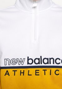 Biała i żółta kurtka sportowa z napisem "new balance ATHLETIC" oraz zamkiem błyskawicznym z przodu.