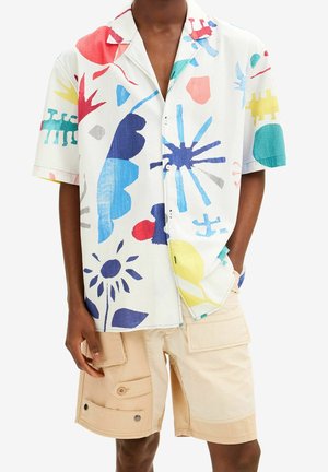Mężczyzna noszący białą koszulę z krótkim rękawem z kolorowymi, abstrakcyjnymi kwiatowymi nadrukami oraz beżowe szorty typu cargo z wieloma kieszeniami.