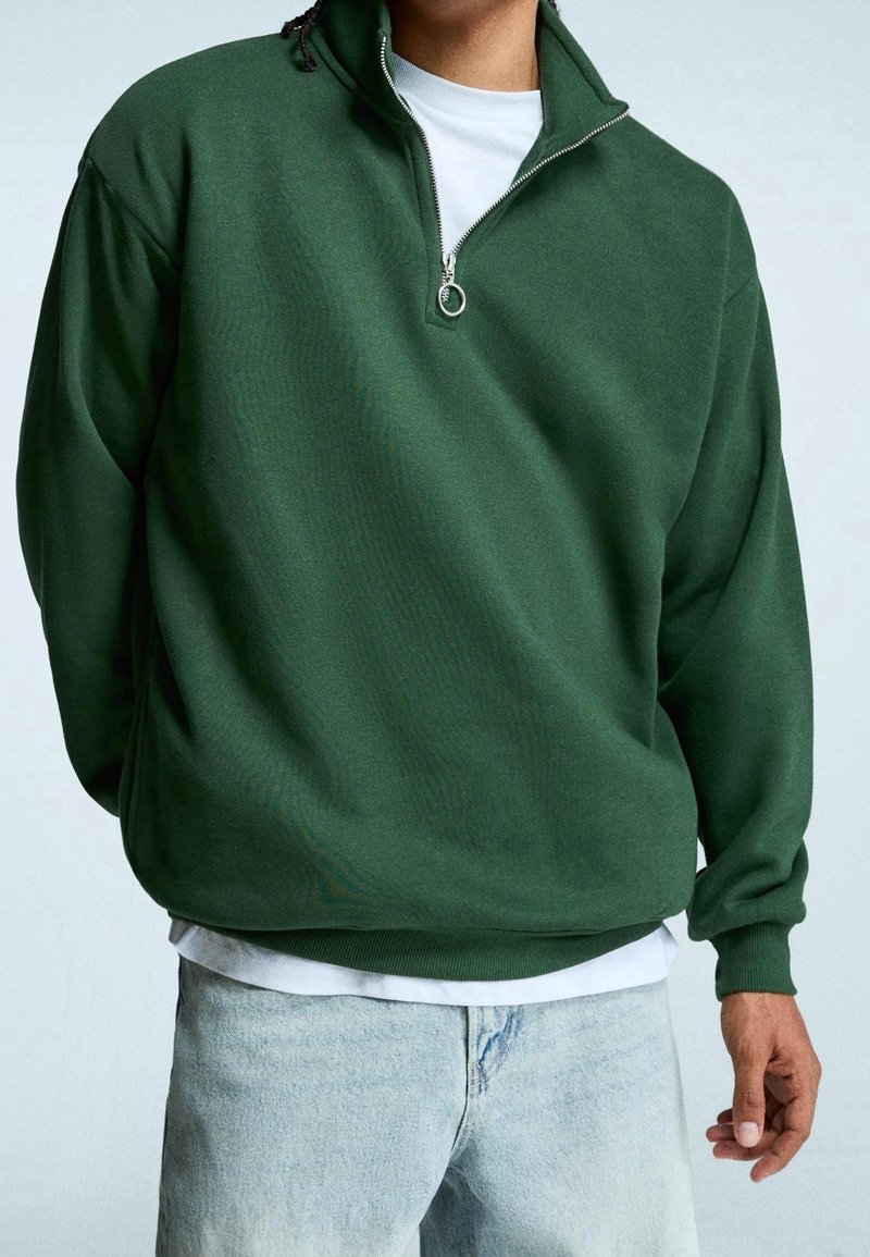 Personne portant un pull à fermeture éclair quart vert foncé par-dessus une chemise blanche et un jean bleu clair, debout avec une main relâchée le long du corps.