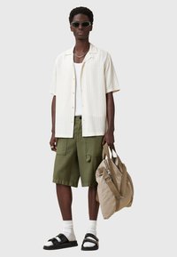 Camicia a maniche corte a quadri crema sopra una canottiera bianca, pantaloni cargo verde oliva, sandali neri e una borsa beige con cerniera e tracolle.