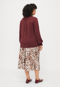 Vínová blůza s dlouhým rukávem a texturovanou tkaninou, doplněná krémově hnědou sukní s květinovým vzorem. Outfit dokončují burgundské baleríny.