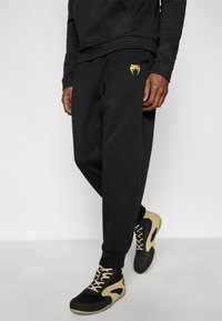 Μαύρο παντελόνι φόρμας από απαλό ύφασμα, με χρυσό λογότυπο στο μηρό και ελαστικές μανσέτες, συνδυασμένο με μαύρα και χρυσά αθλητικά παπούτσια.