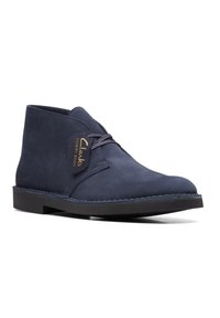 Botas desérticas de ante azul marino con punta redonda, diseño de tobillo bajo, cierre de cordones y suela de goma negra. Acento de logotipo dorado en el lateral.