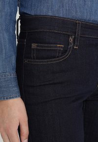 Los jeans de mezclilla azul oscuro presentan un corte ajustado, con un diseño tradicional de cinco bolsillos y costuras amarillas contrastantes en las costuras.