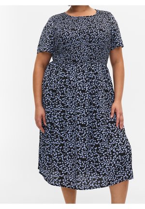 Vrouw die een zwarte midi-jurk draagt met korte mouwen en een kleine blauwe bloemenprint, staand met handen ontspannen langs het lichaam.
