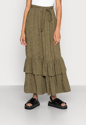Femme portant une jupe à volants à motif vert olive avec une ceinture à la taille, un haut blanc et des sandales à plateforme noires sur un fond clair.
