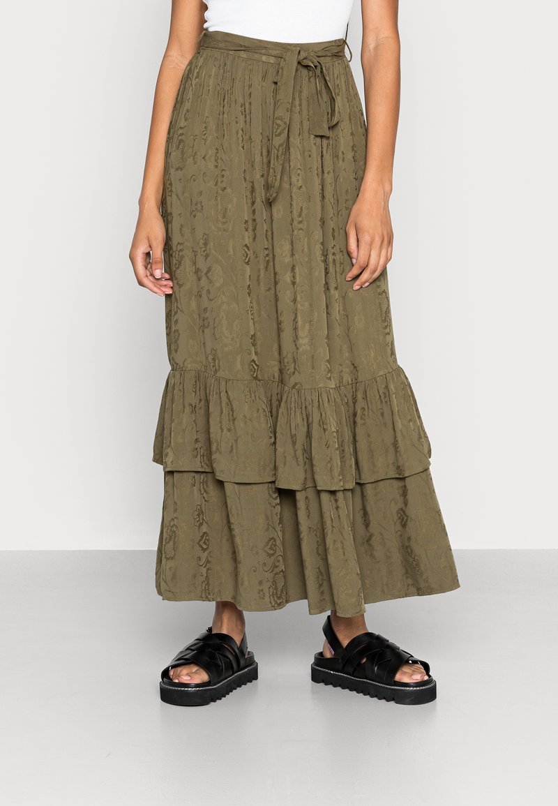 Femme portant une jupe à volants à motif vert olive avec une ceinture à la taille, un haut blanc et des sandales à plateforme noires sur un fond clair.