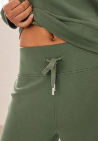 Green jogger pants made of soft material, featuring a drawstring waist with a textured tie and small silver accents on the ends.