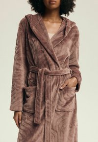 Burgundowy pluszowy szlafrok z kapturem, ozdobiony teksturowanymi falistymi wzorami, z paskiem w talii i dwoma przednimi kieszeniami. Miękka, gruba tkanina.