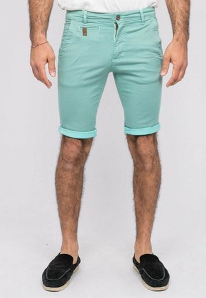 Homme portant un short vert menthe roulé, une chemise blanche, des chaussures noires sans lacets, debout devant un fond uni.