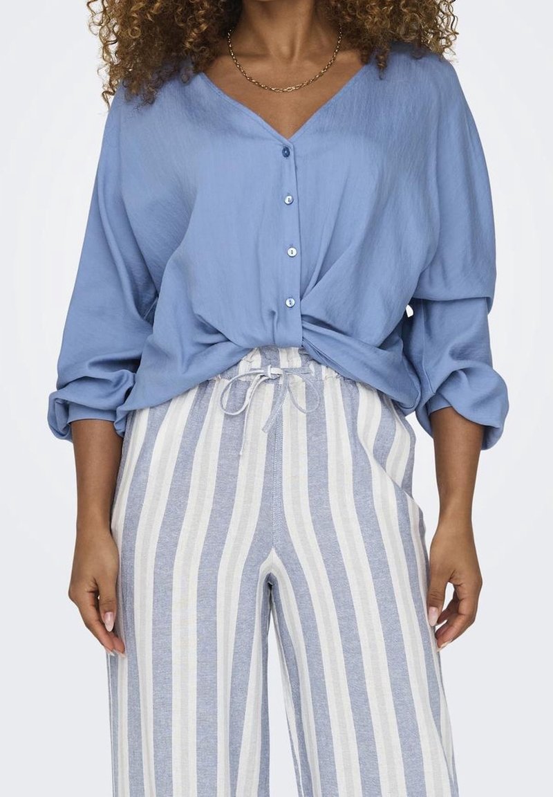 Ljust blå knapparblus med V-ringning och knuten fram, kombinerad med randiga vida byxor i blå och krämfärgade toner. Mjuk tygtextur.